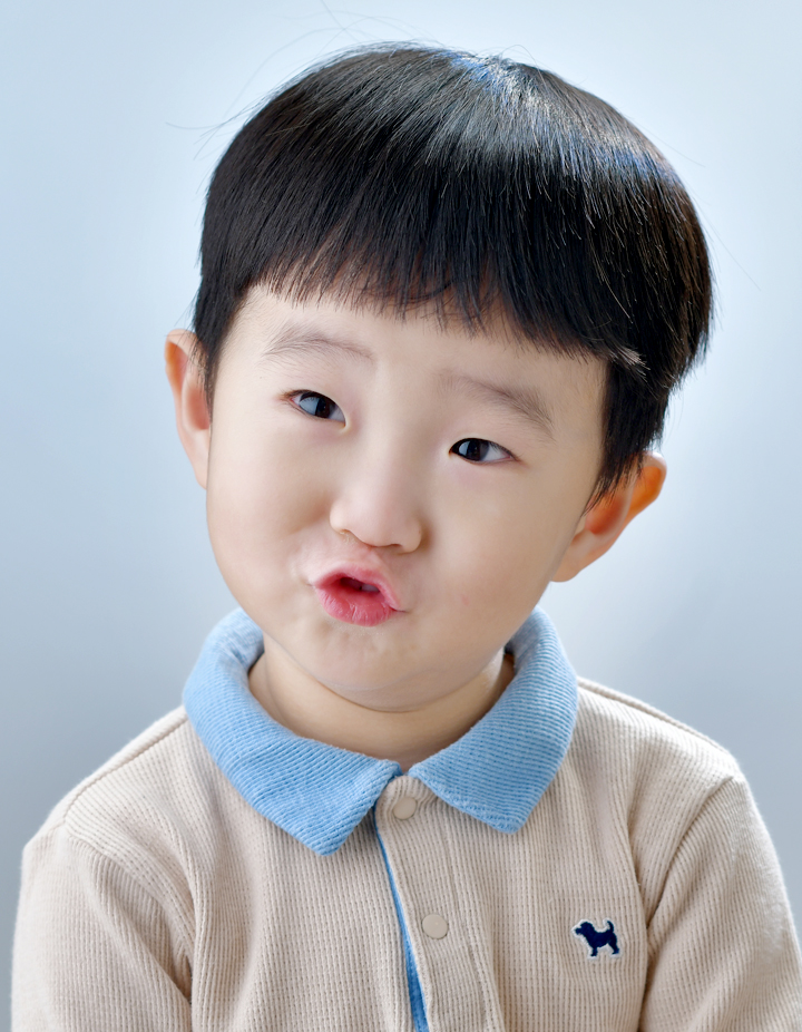 본문 사밝은 하늘색 배경에서 파란색 카라가 있는 베이지색 니트를 입고 촬영한 남자아이 프로필사진. 짧은 머리에 입술을 동그랗게 내민 장난스러운 표정이 돋보이는 어린이 증명사진.진 2