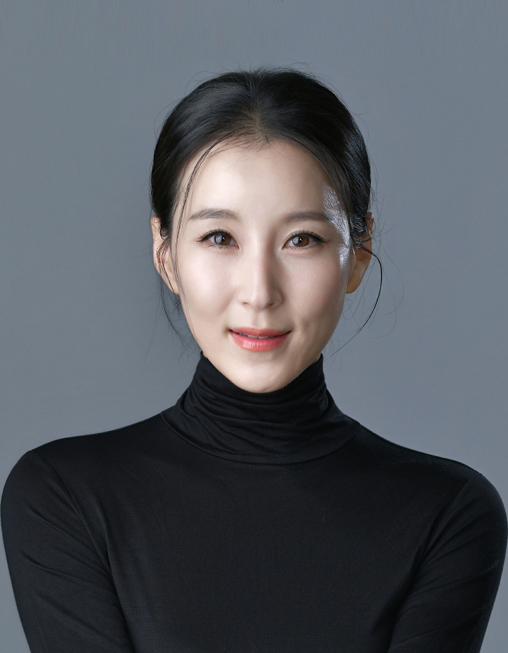 검은색 터틀넥을 입은 여성의 증명사진. 회색 배경에서 단정하게 묶은 헤어스타일과 은은한 미소로 차분하고 세련된 분위기를 표현한 인물사진.